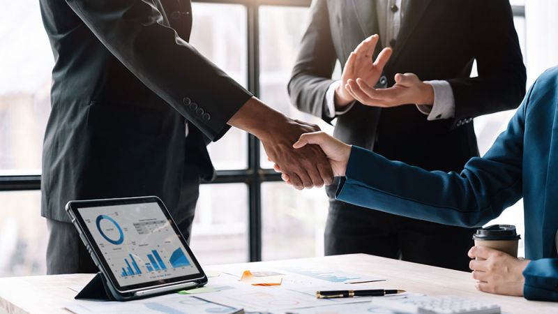 Two professionals shake hands across a desk, signifying a business agreement or partnership.
